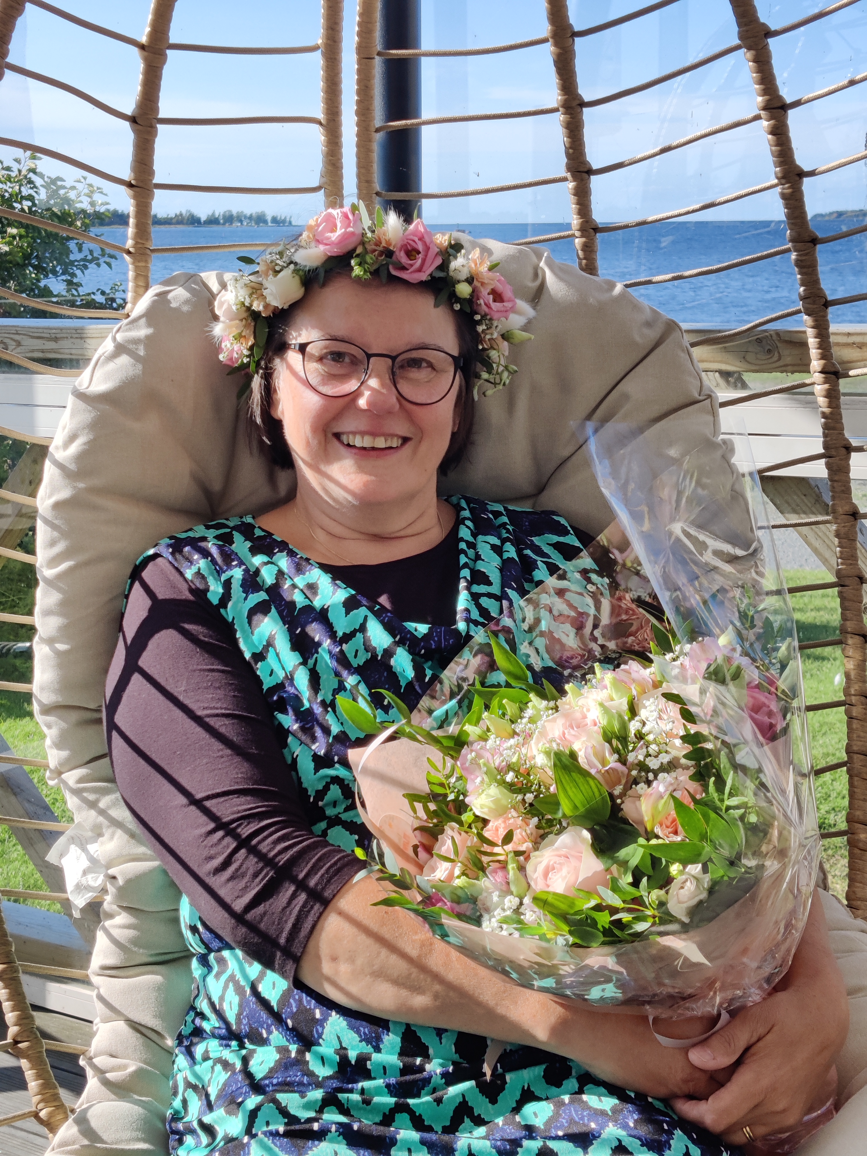 Iloisesti hymyilevä nainen istuu korituolissa kukkaseppele päässään ja kaunis kimppy sylissään.